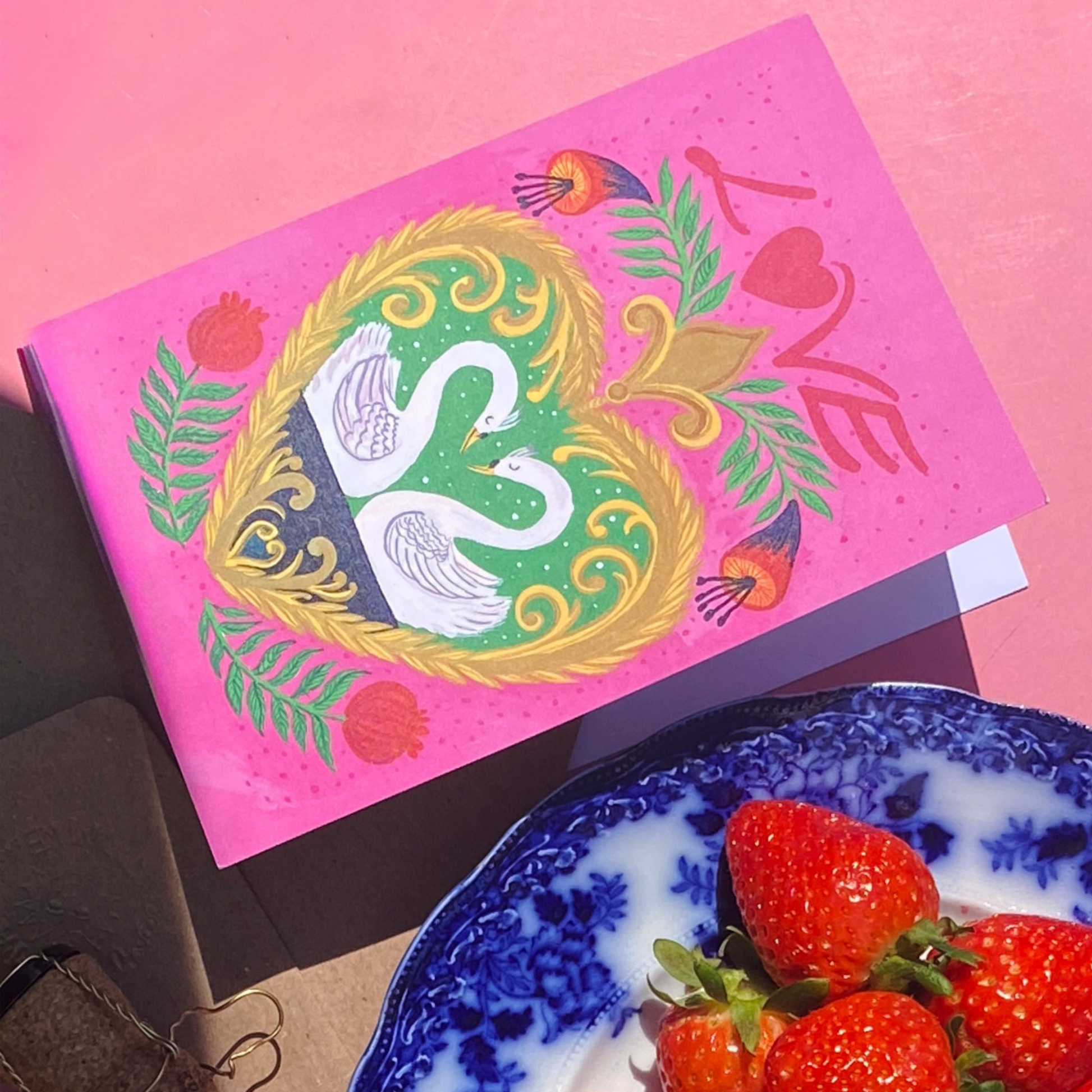 Decorative card with a swan design on a pink background, next to strawberries on a blue and white plate.