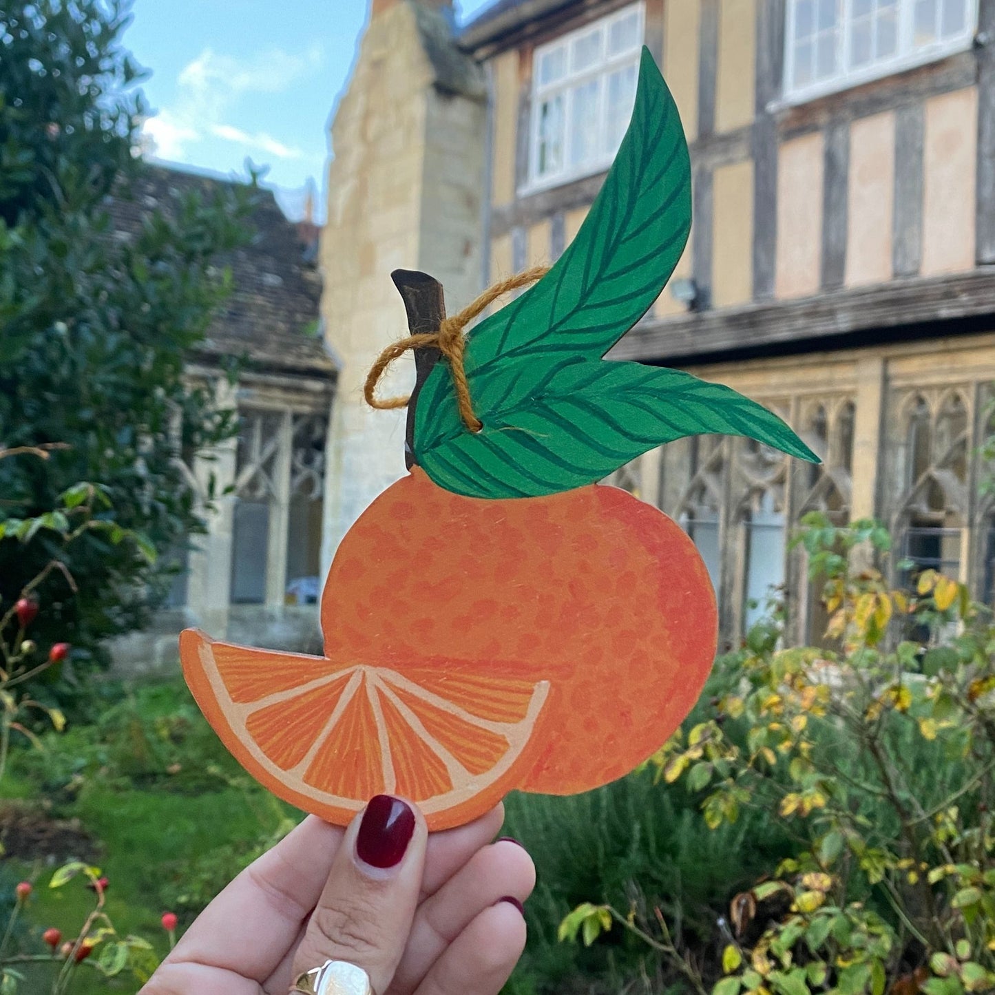 Hand holding a paper cutout of an orange with green leaves in front of a building.