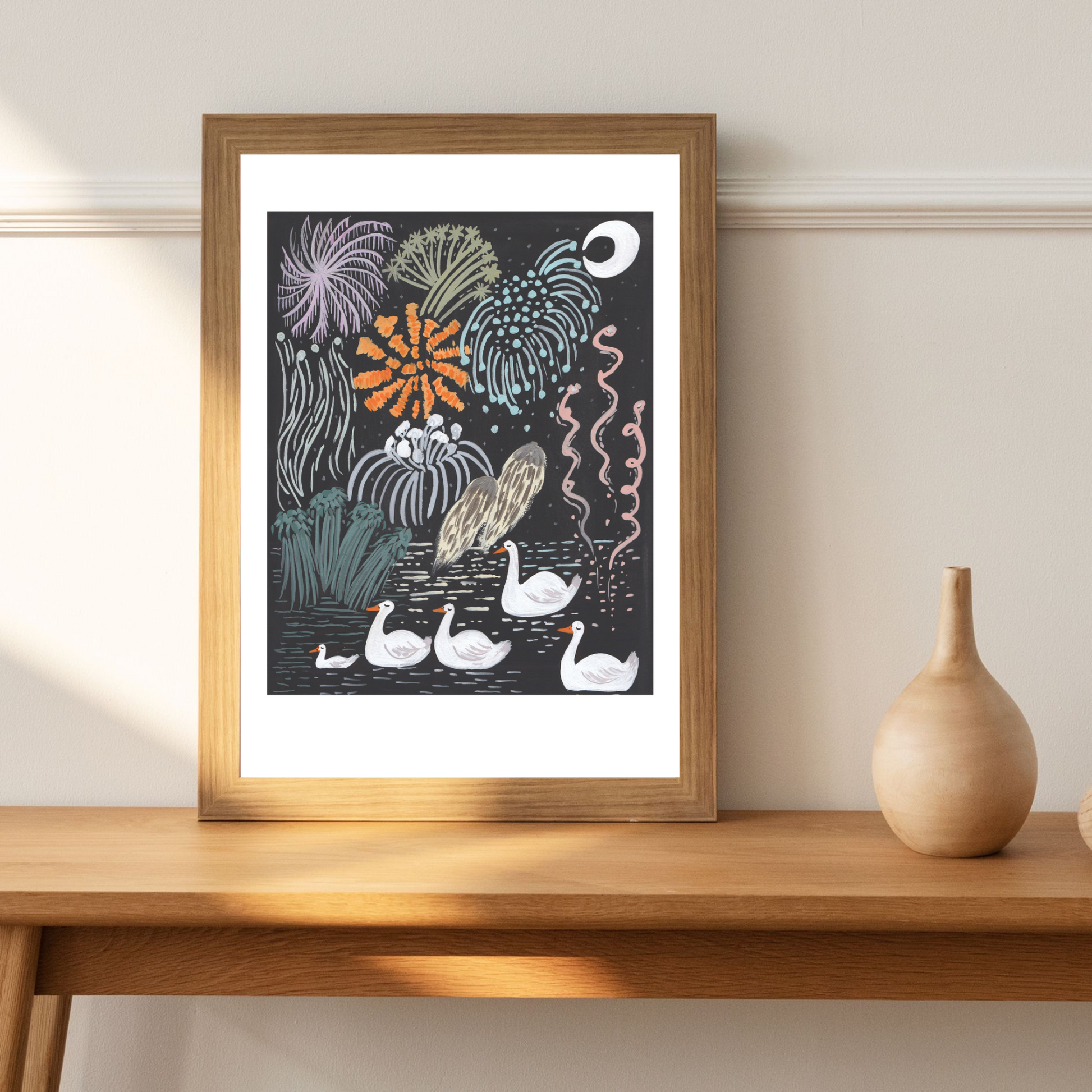 Framed artwork with swans and fireworks on a wooden table.