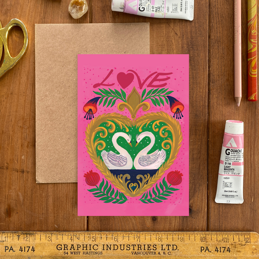 Colorful card with swans and 'LOVE' on a wooden surface with art supplies.