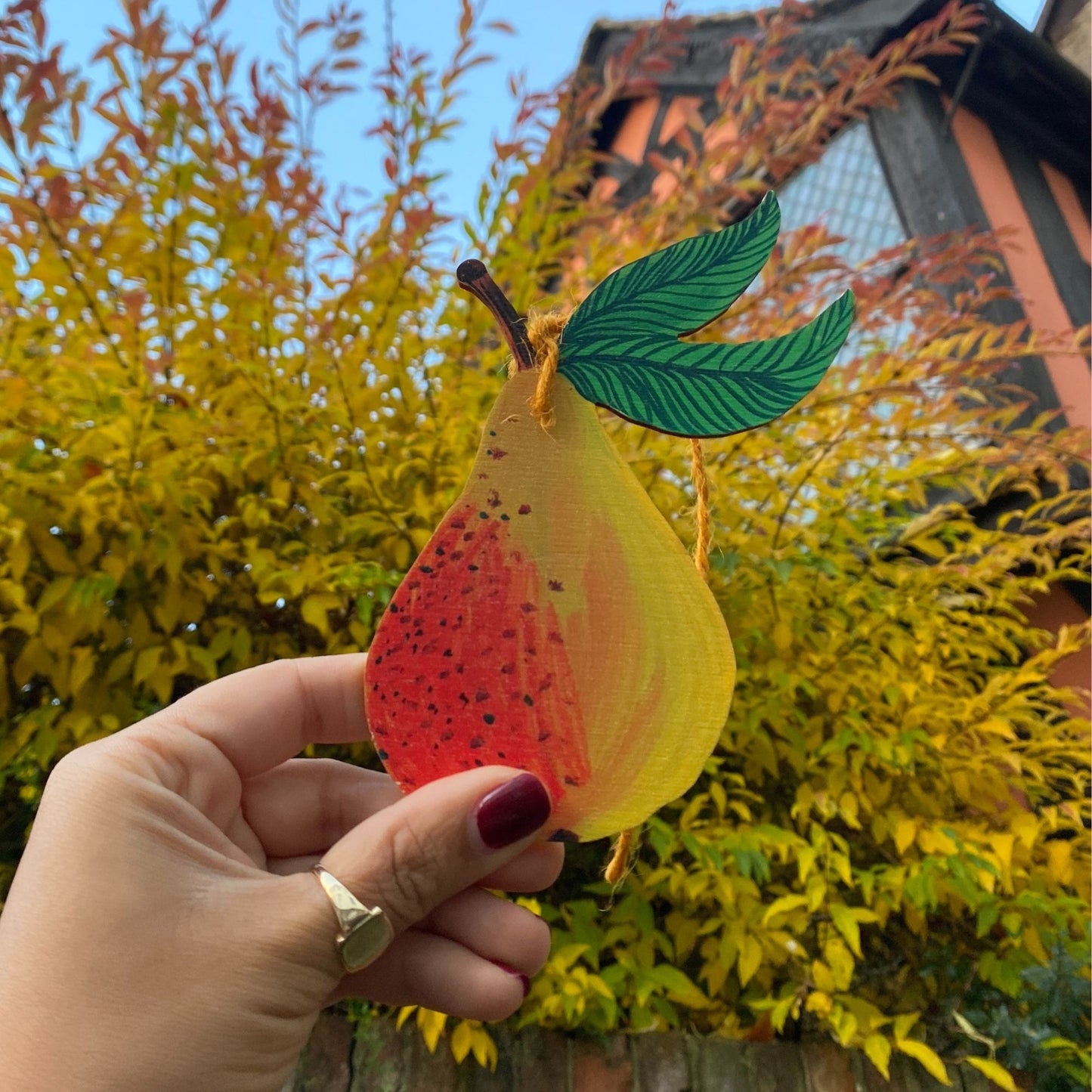 Hand holding a colorful leaf-shaped object with a blurred background of trees and a building.