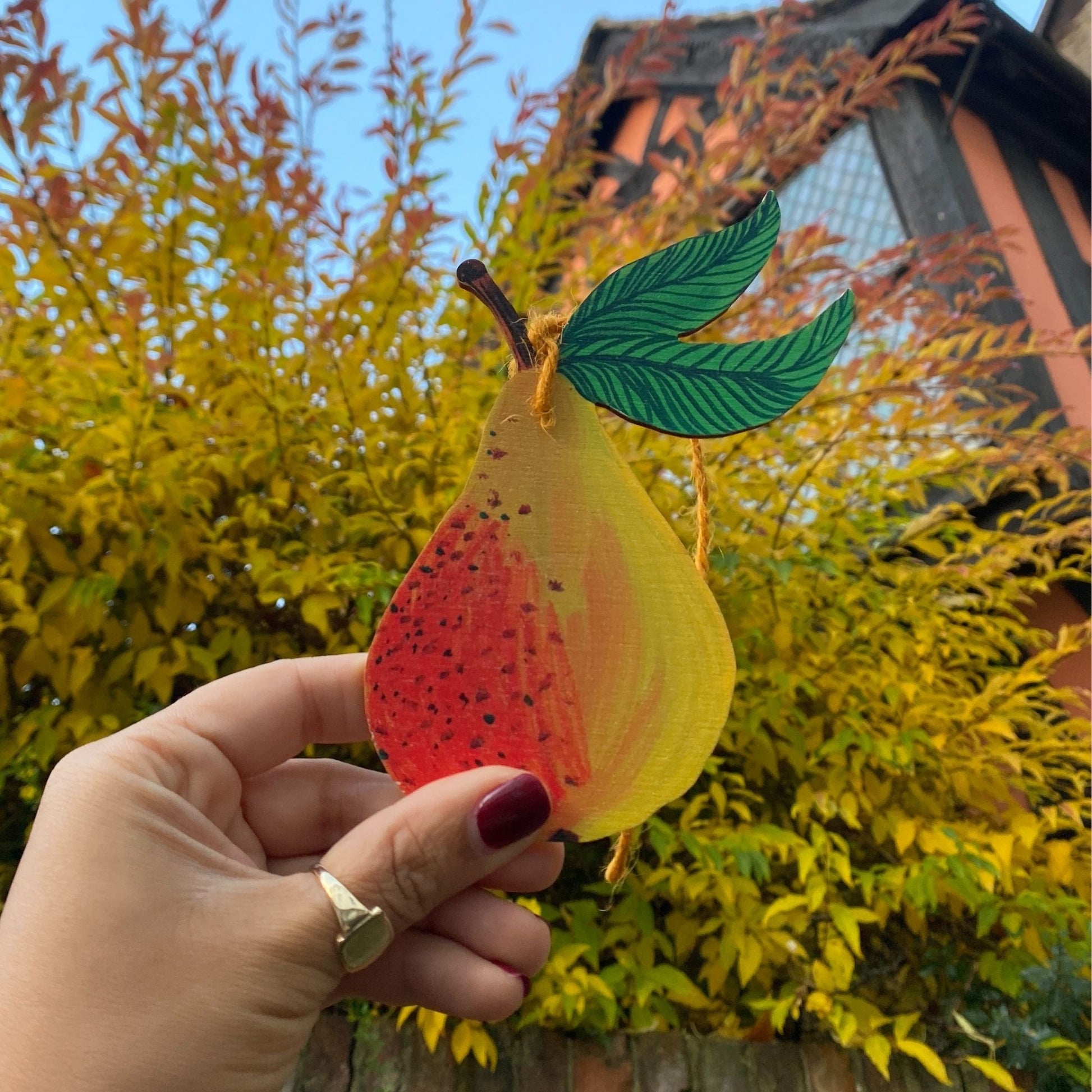 Hand holding a colorful leaf-shaped object with a blurred background of trees and a building.