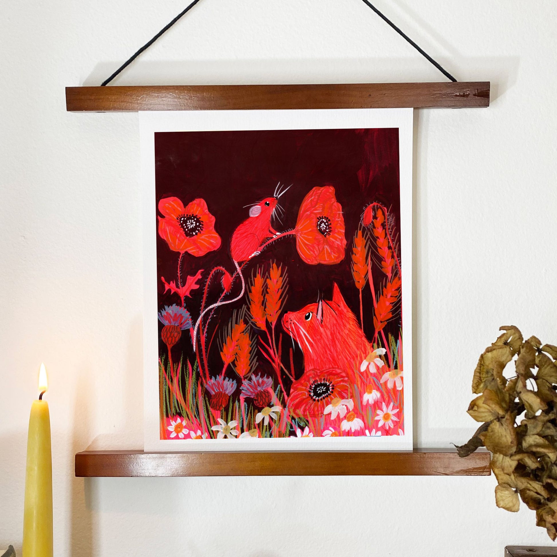 Framed artwork of red frogs and flowers on a white wall with a candle and plant.