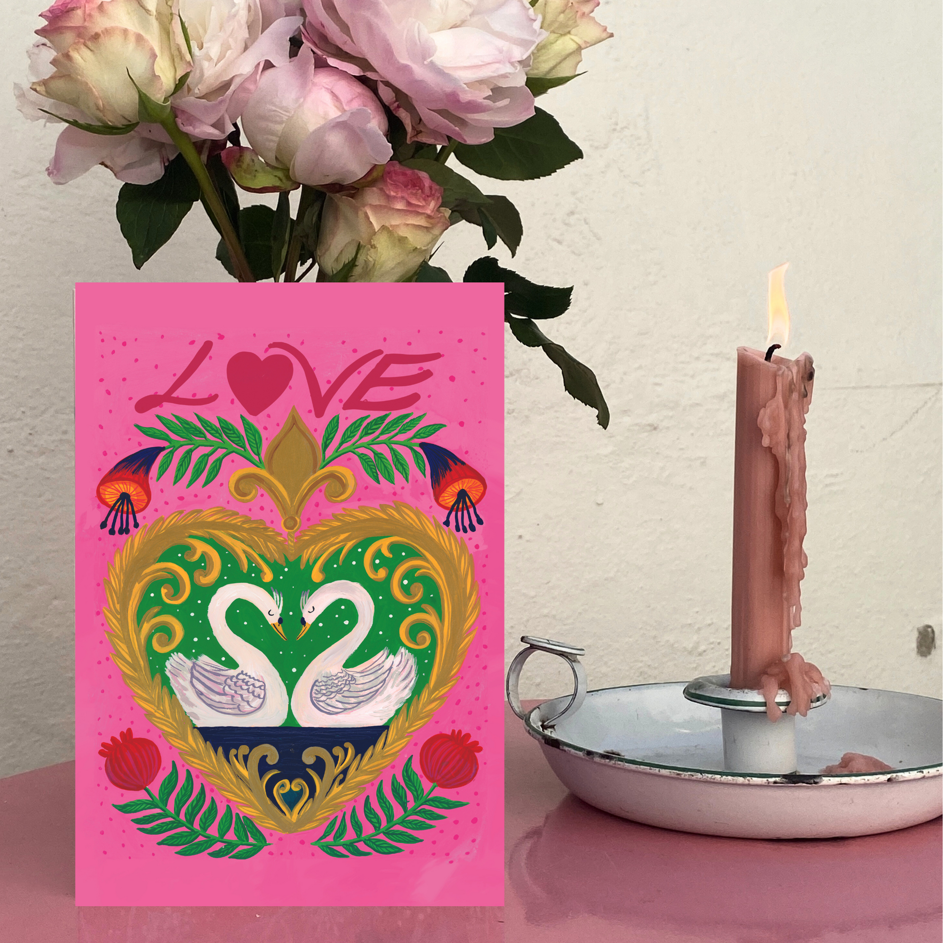 Pink card with decorative heart and swans design next to a lit candle and flowers.