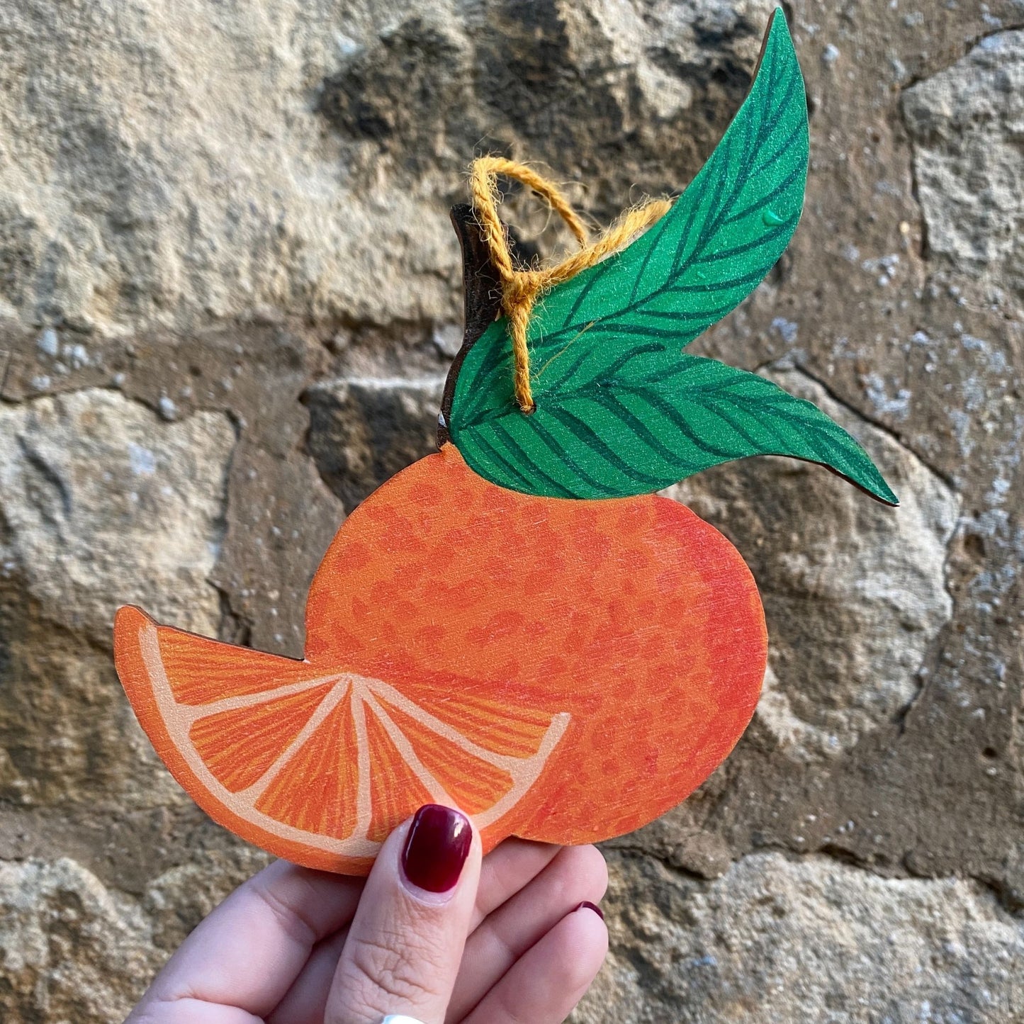 Hand holding a paper cutout of an orange with leaves against a stone wall background