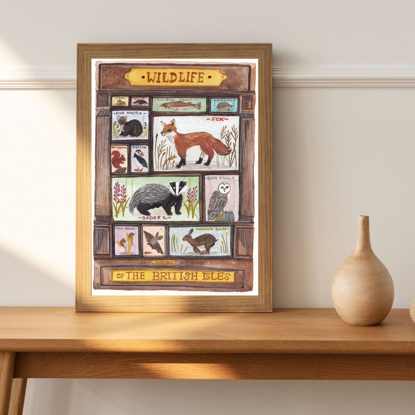 Framed artwork of British wildlife on a wooden table with a vase.
