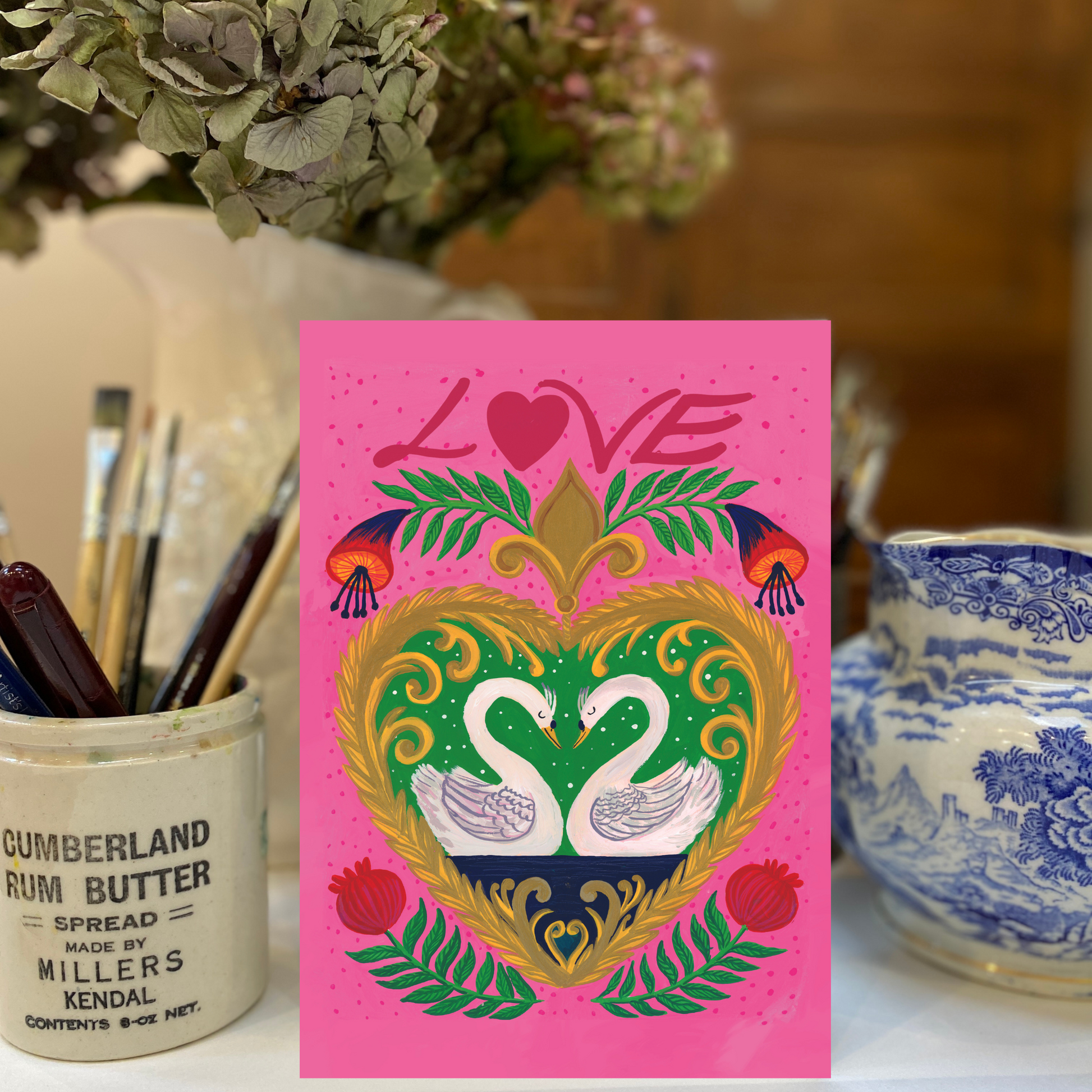 Pink card with decorative heart design featuring swans, surrounded by art supplies and a vase.