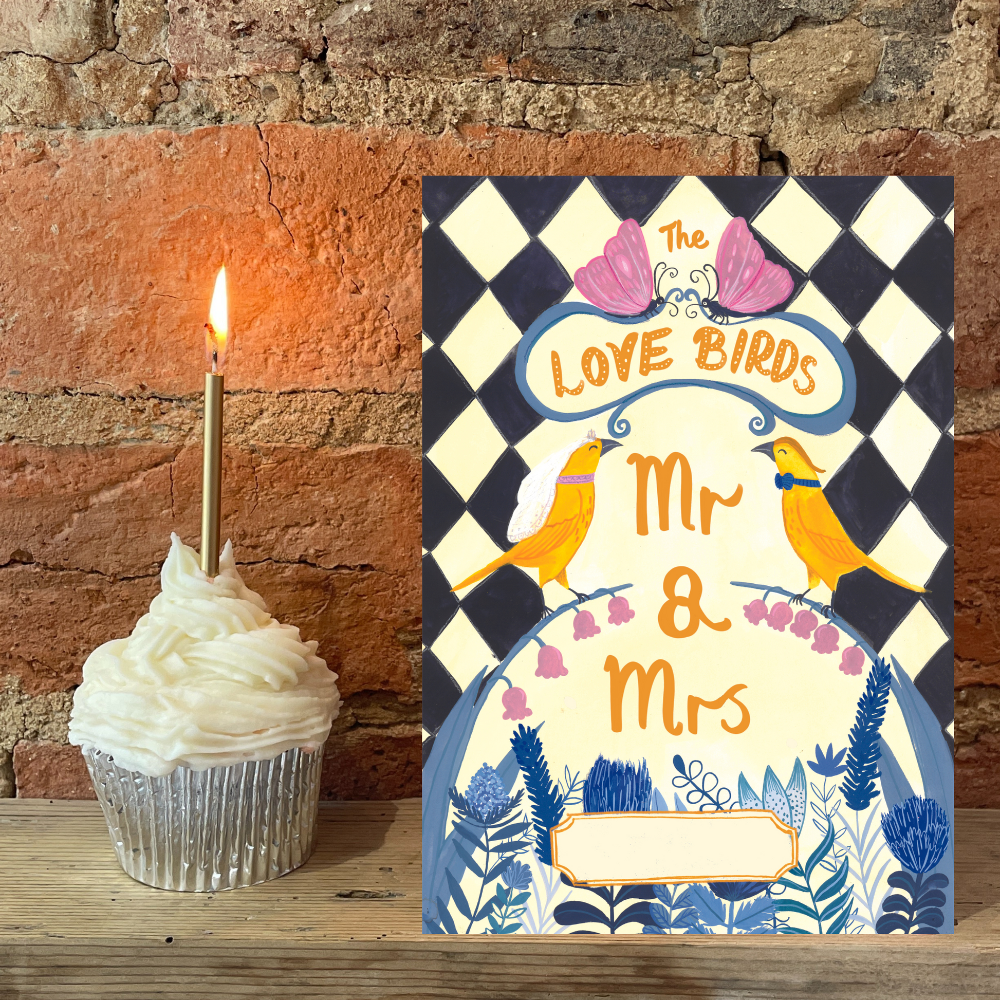 Decorative card with 'Mr & Mrs' text and two birds, next to a cupcake with a lit candle against a stone wall background.