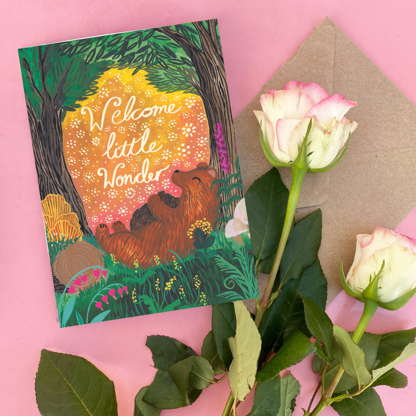 Greeting card with bear illustration and 'Welcome little wonder' text, surrounded by flowers and an envelope on a pink background.