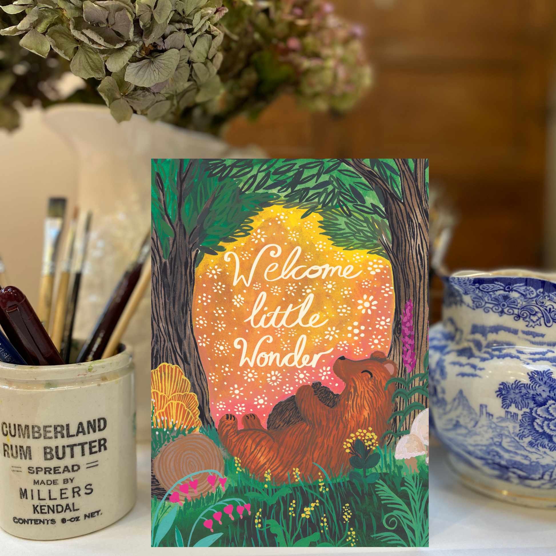 Card with bear and floral design labeled 'Welcome little wonder' on a table with decorative items.